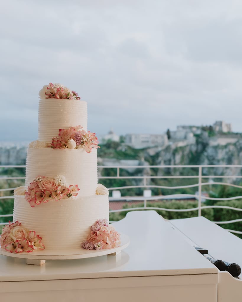 Torta nuziale a tre piani con fiori rosa su un pianoforte bianco, sulla terrazza di Villa Hera; alle spalle il Castello di Milazzo.